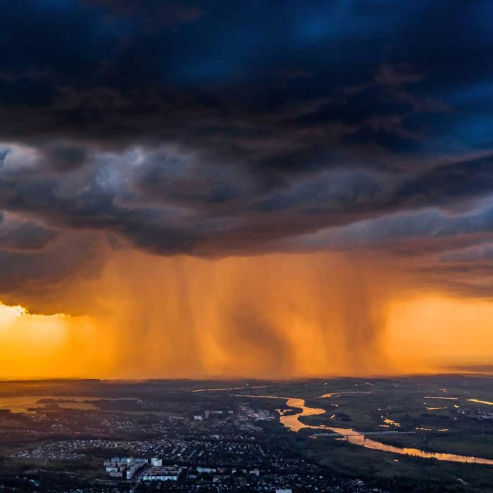 Dramatische Gewitterwolken mit starkem Regen über einer Landschaft bei Sonnenuntergang, durchzogen von Flüssen und Feldern.