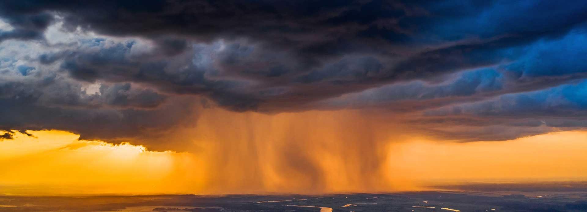 Dramatische Gewitterwolken mit starkem Regen über einer Landschaft bei Sonnenuntergang, durchzogen von Flüssen und Feldern.